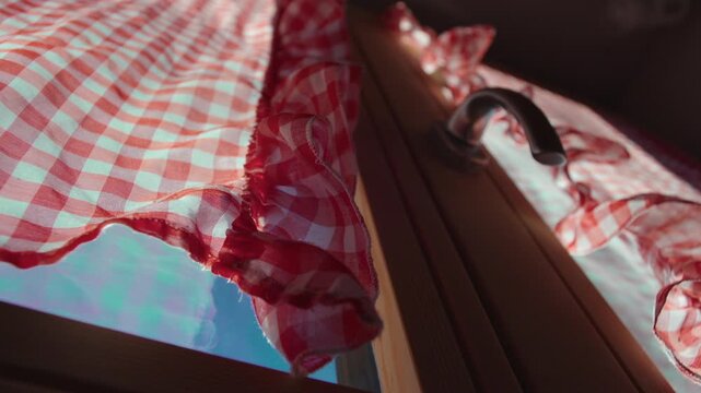 Red and white checkered curtain hanging over chalet window, sunlight passing through fabric beside wooden frame and metal handle