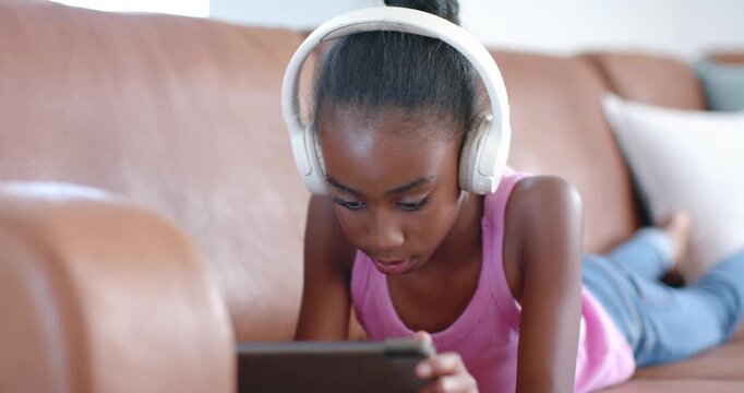 African American teen girl propping on elbows, tapping tablet while wearing headset on sofa at home