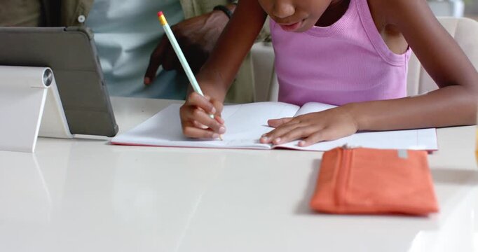 African American man supervising girl child writing in notebook at table, tablet guiding work
