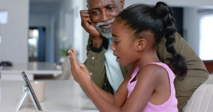 African American grandpa cheering youth female learning numbers after tablet cue at kitchen counter