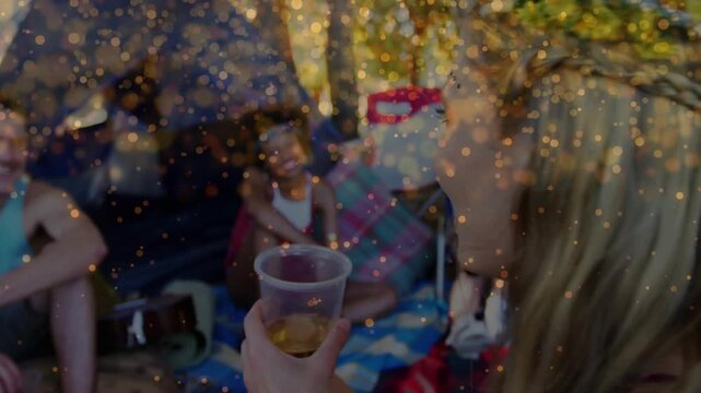 Camera panning revealing woman lifting plastic cup to drink at campsite while golden bokeh floating