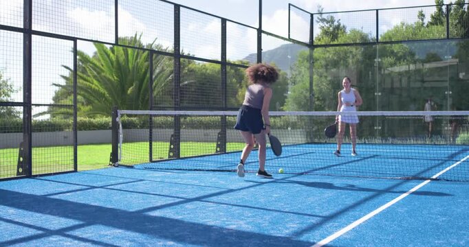 Diverse female pair rallying after serve, returning shot on padel court, player lunging with paddle