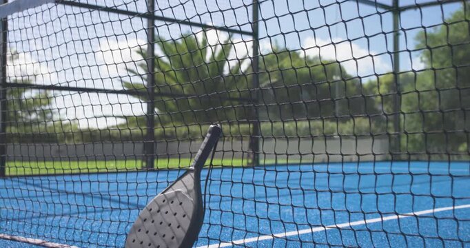 Tilting-pulling camera is revealing paddle leaning against net and green ball on court to showcase