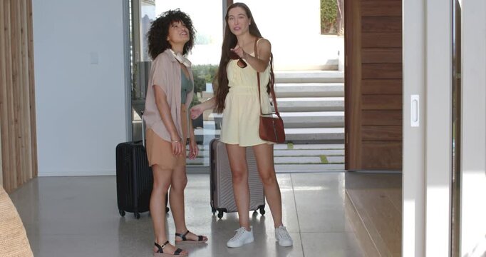 Diverse female friends arriving at entry, pointing and smiling, showing door feature with luggage