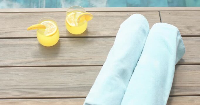 Two rolled light-blue towels staying on deck as camera is pulling back, revealing pool and drinks