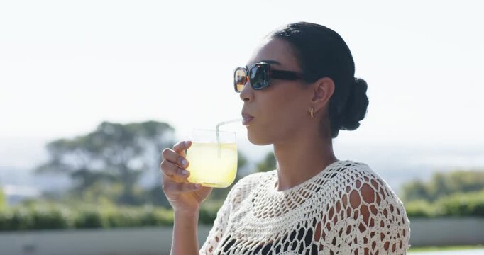 African adult woman thirsty, raising sipping yellow drink via straw on patio in shades, copy space
