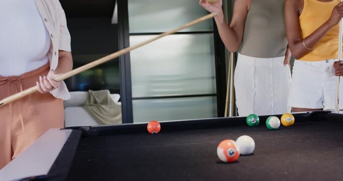 Diverse female friends playing at black pool table, shooter aiming and striking cue ball, cheering