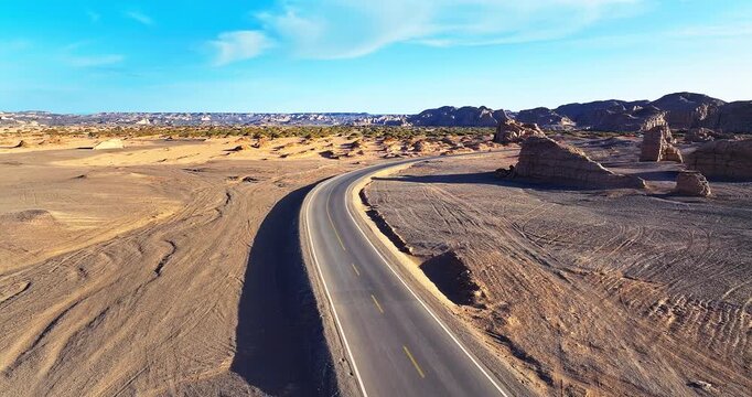 Aerial view of a winding asphalt road through the Yardang landforms in Xinjiang, China. Desert highway and unique rock formations under a clear blue sky.