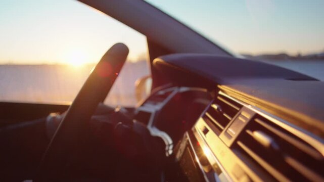 Car interior dashboard and steering wheel illuminated by winter sunset, driver perspective facing snowy countryside through windshield during evening drive