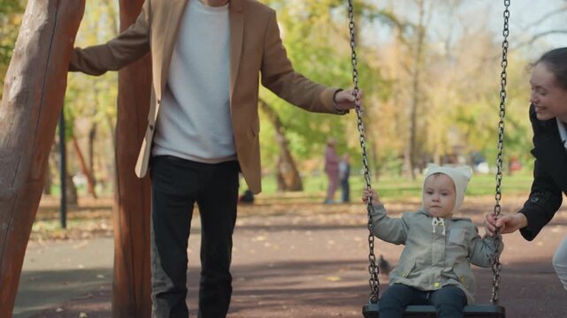 Closeup white toddler gripping swing chain, park autumn backdrop with father steadying frame and mother holding opposite side, attentive gaze and protective hands, marriage partnership expressed