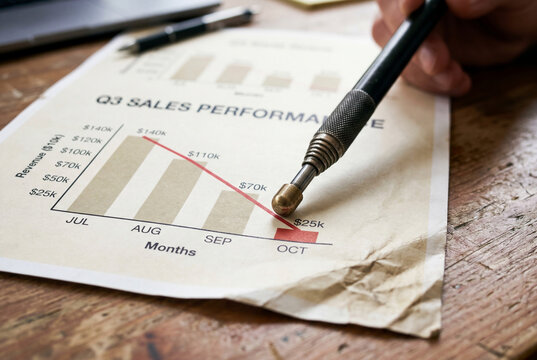 Macro shot of a hand pointing a technical pen to a declining sales performance graph on wrinkled paper, representing financial crisis and failure
