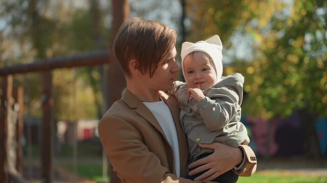 Caring father in autumn park with infant. Tender moment between dad and infant in sunlit park. Intimate family scene showing father adjusting hat on smiling baby during autumn afternoon