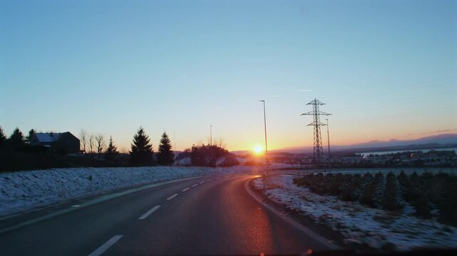 Winter highway curving toward sunrise over snow covered countryside and distant mountains, golden dawn light illuminating rural landscape