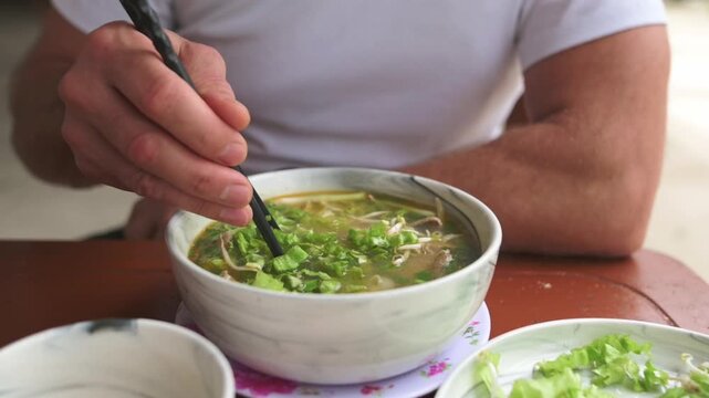 Caucasian Adventurer Lifting Noodles From Vietnamese Pho Bo, Slow Slurp Action With Chopsticks, Steam Rising From Bowl, Fresh Herbs Clinging To Noodles, Satisfying First Bite Captured