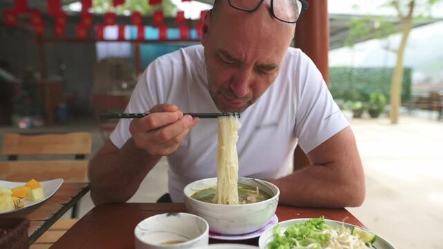 White Man Outdoor Table Eating Pho Bo, Slurping Hot Noodles With Chopsticks, Glasses On Head, Fresh Herbs And Lime On Wooden Table, Rustic Openair Vietnamese Cafe, Steam Rising, Enthusiastic