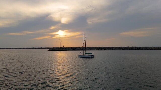 4K peaceful drone shot of a lone yacht resting on calm ocean water by a harbor pier at dusk