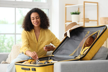 Young African-American woman with mobile phone packing suitcase for trip on sofa at home