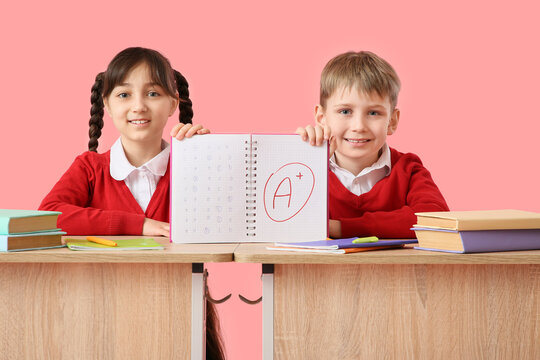 Happy little pupils holding notebook good results of school tests at desk on pink background