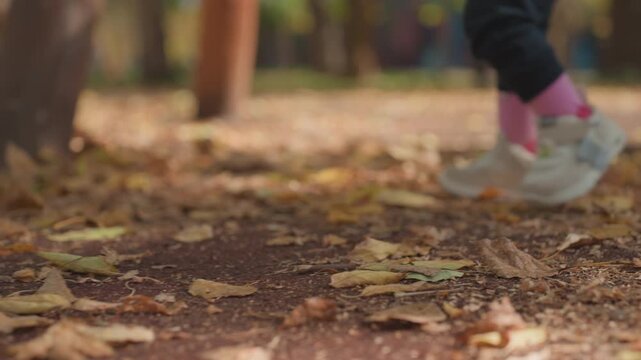 Child stumbling then exploring ground among fallen leaves, closeup shoes and pink socks, gentle adult legs nearby offering support, autumn light and tactile discovery, candid family play and learning