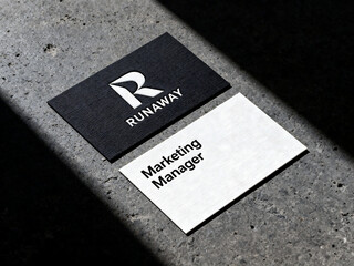 A pair of modern business cards, one black with a white logo and "RUNAWAY", the other white with "Marketing Manager", on a textured concrete surface with strong shadows.