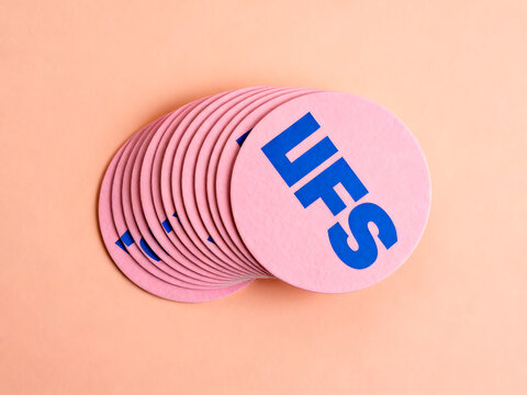 A stack of round pink coasters with blue "UFS" lettering, fanned out on a peach background.
