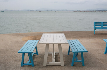Serene coastal spot featuring a white wooden picnic table and blue benches, perfectly inviting someone to sit back, relax, and soak in the tranquil ocean view on a calm, overcast day.