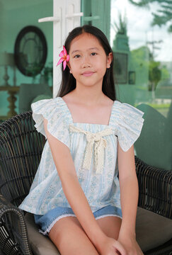 Beautiful young Asian girl with pink plumeria flower in her hair is smiling widely while sitting comfortably in an outdoor wicker chair.