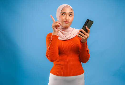 Thoughtful young Muslim woman holding a smart phone while pointing to the copy space upward.