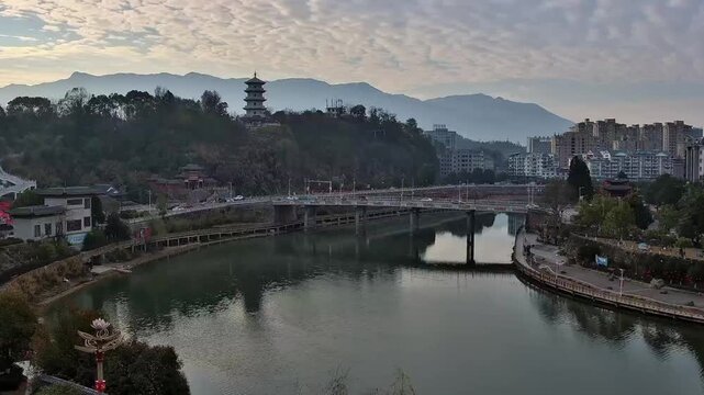 A peaceful morning view of a town in Baoshan, Yunnan, China. The scene features a traditional Chinese pagoda on a hill, a modern bridge across the river, and distant mountains under a cloudy sky.