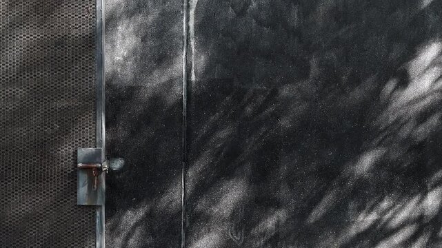 Elegant black terrazzo stone wall beside industrial metal gate with shimmering sunlight and moving tree shadows across surface. Modern architectural background blending urban texture, luxury stone.