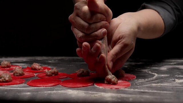 Closeup of chef hands piping meat filling onto circular red pasta dough discs on floured dark surface in low light culinary preparation sequence
