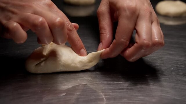 Closeup hands shaping soft dough on metal countertop, artisan preparation of handmade dumplings or buns in low light kitchen cooking process sequence