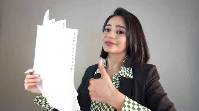 Closeup portrait of happy smiling beautiful young Indian business woman manager boss hold project document papers looking at camera showing thumb up hand gesture, over grey studio background.