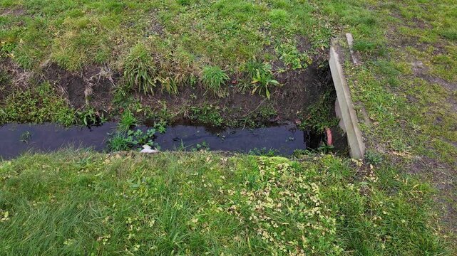 Concrete culvert under a path and drainage ditch in a grassy field, rainwater management system in rural area, hydrological infrastructure and plot drainage in close-up view.