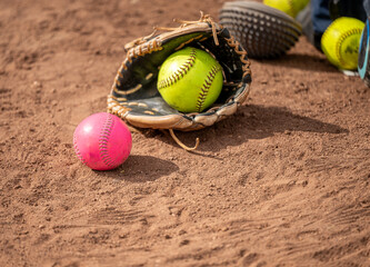 Obraz premium Close-up of a leather softball glove holding a yellow ball next to a pink softball on a dirt field.