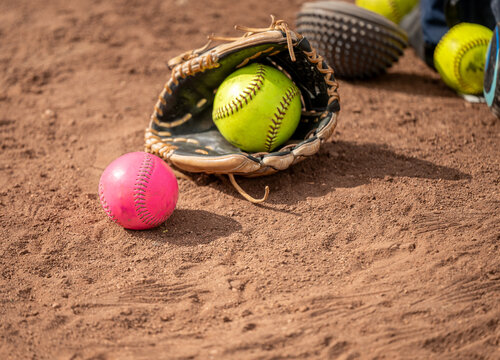 Close-up of a leather softball glove holding a yellow ball next to a pink softball on a dirt field.