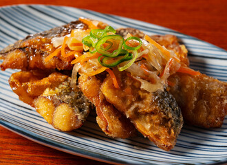 Fototapeta premium Close-up of Japanese deep-fried mackerel with sweet and sour vinegar sauce and vegetables on a striped plate over a wooden table