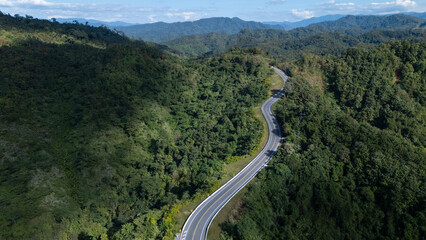 A winding road cuts through lush, green mountainous forest under a partly cloudy sky.