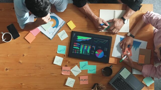 Top aerial view of diverse team writing colorful sticky notes to brainstorm marketing idea while looking at financial chart. Multicultural smart business people taking a note about plan. Convocation.