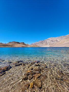 Wide landscape view of the pristine transparent blue waters of Chandratal Lake revealing underwater stones in the high altitude Himalayas
