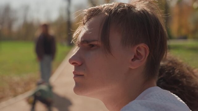 White young man in park looking contemplative on bench, blurred figure walking along pathway in background, autumn leaves and warm sunlight, cinematic shallow depth, candid portrait capturing