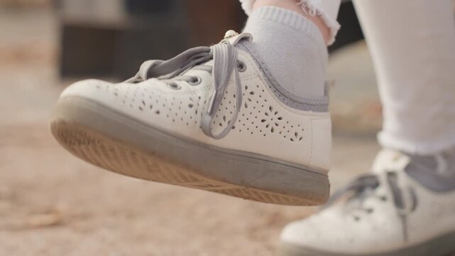White sneaker closeup on child swinging foot at playground capturing family warmth and playful innocence sock and lace detail, worn sole on dusty ground, candid movement, leisure mood suggesting