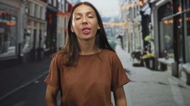 Young hispanic brunette woman sticking out tongue with hands framing face on a cobbled city street wearing a brown shirt; playful mischief.