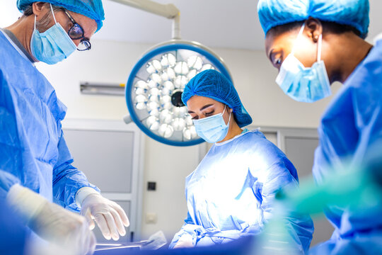Group of surgeons performing surgery in operating room. Team of medical specialists focused on procedure under bright theater lights. Healthcare and medicine concept. Doctors in blue sterile scrubs.