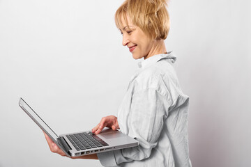 Smiling woman with short blonde hair using laptop computer while standing against white background. Concept of technology, remote work, communication, and digital lifestyle.