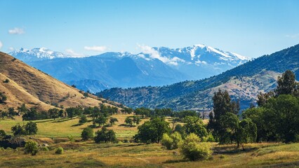 Snow-capped Sierra Nevada peaks rise above rolling California foothills dotted with oak trees and open grassland under a clear blue sky. © The Hiker's Eye
