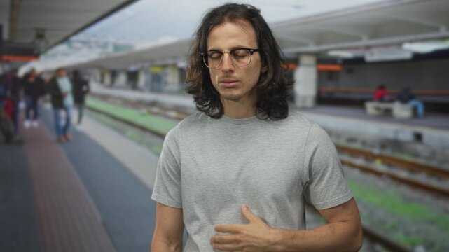 Man with long hair and glasses clutch chest, hand over sternum suggesting heartattack on train platform building near railway tracks; anxiety urgency.