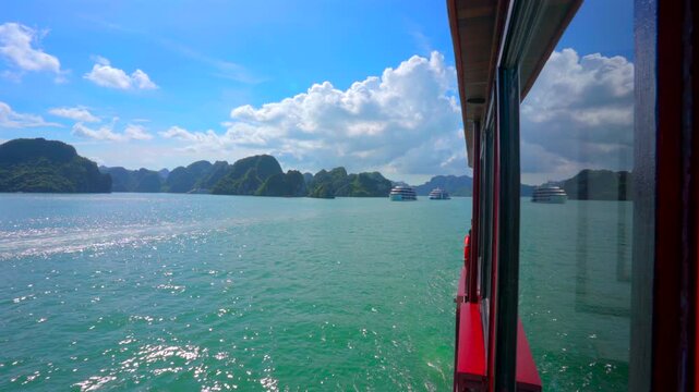 A bright side-view from a red cruise ship, showing the turquoise sea and karst islands reflecting in the cabin window under a brilliant blue sky with white clouds.