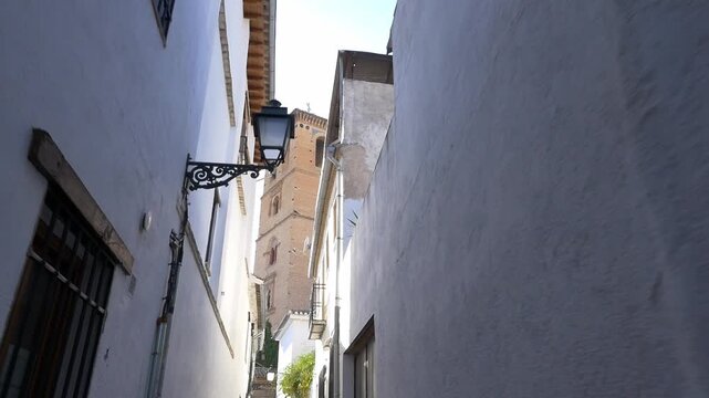 POV walking shot through a narrow alley in the Albaicin district, revealing the Mudejar tower of San Bartolome Church. Bright daylight, white facades, and traditional Spanish atmosphere.