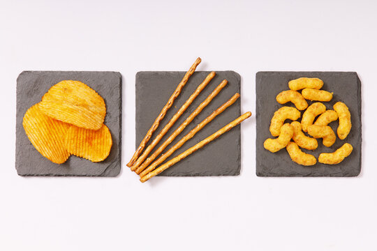 Salzgeb&auml;ck, Kartoffelchips, Erdnussflips und Salzstangen auf quadratischen Schieferplatten